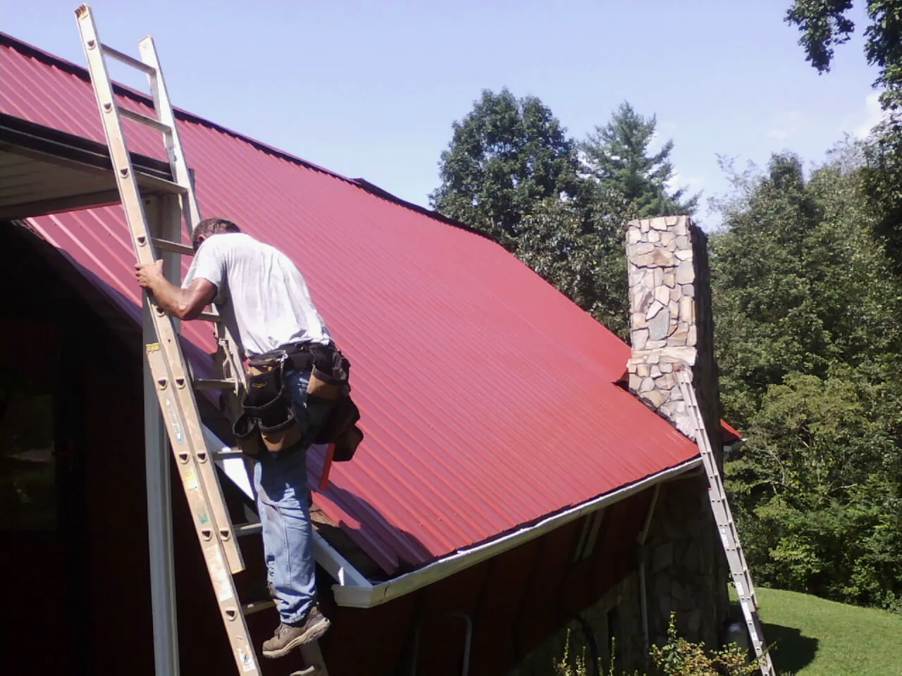 Roofer on a ladder beside a red metal roof and stone chimney — Soffit & Fascia Repair by Roofing Brothers in Lake Harmony